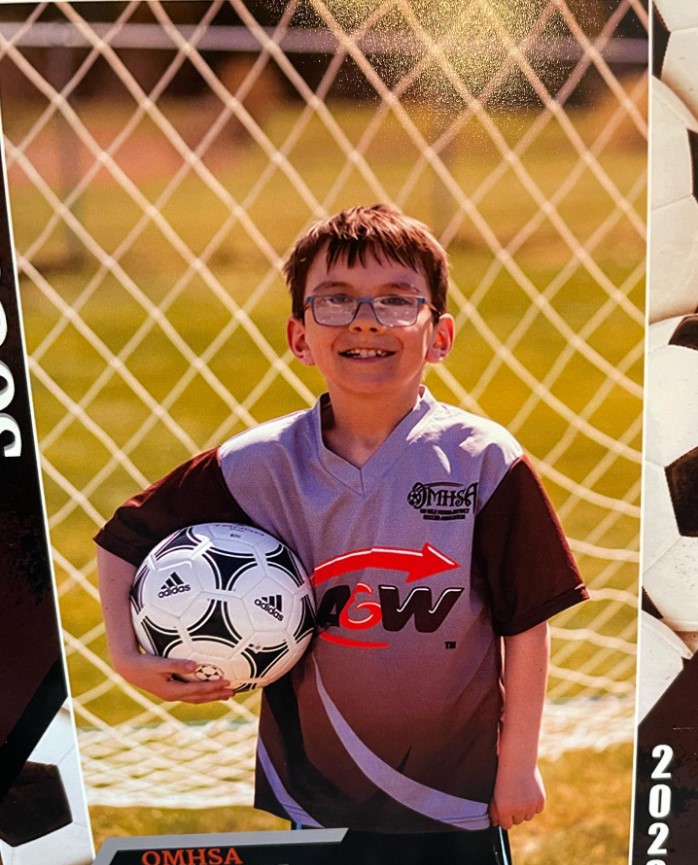 un niño se para frente a una red de fútbol sosteniendo una pelota con una camiseta.  Amber Vigh anunció la muerte de su hijo de nueve años en Facebook