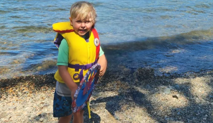 Three-year-old Kash Bakker stands at the water's edge wearing a bright yellow lifejacket. The toddler was identified in a GoFundMe campaign as the child who was killed when a tree fell in the Okanagan Lake Provincial Park campground in late July 2023