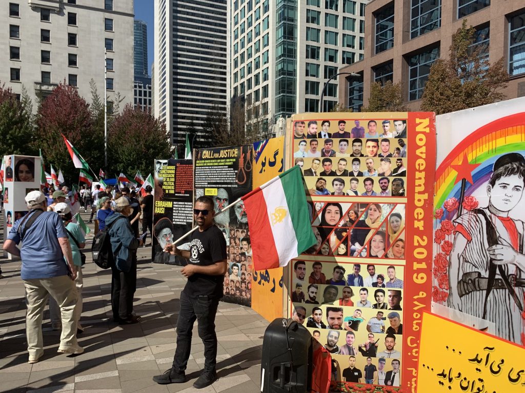 iran-protest-2 A man holds an Iranian flag in front of a large poster filled with the faces of Iranian youth who have been killed in Iran
