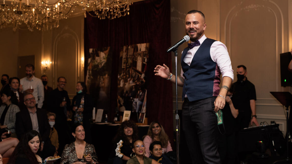 Danny Ramadan stands at a microphone wearing a white shirt and black vets while speaking to a crowd of people at An Evening in Damascus in 2021