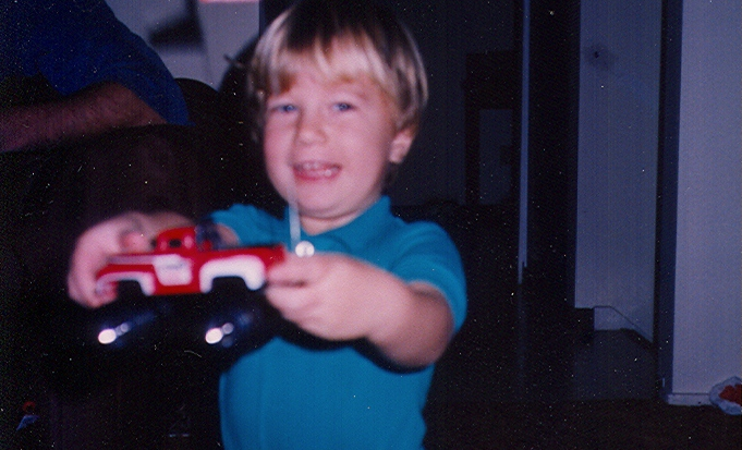 Michael Dunahee, a little boy with blonde hair, wearing blue holds up a red toy truck in front of him