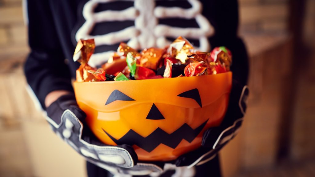 Halloween candy in a bowl in the shape of a Jack-o'-lantern is held by a child wearing a skeleton costume.