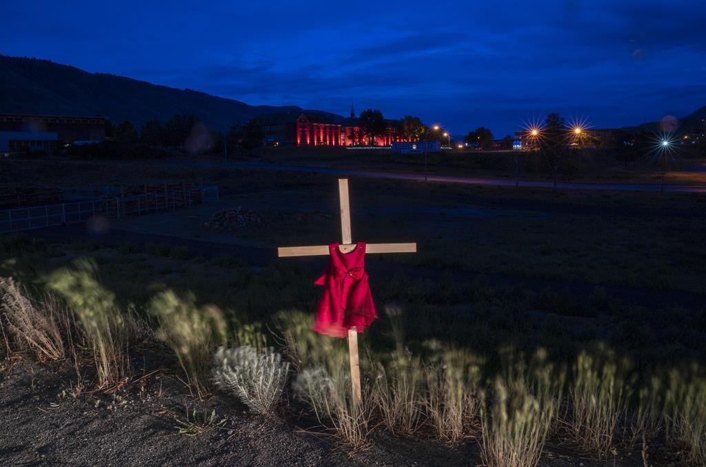 A child's dress is seen on a cross outside a former residential school in Kamloops, B.C., Sunday, June 13, 2021. An interim report from an international group hired to provide advice on identifying and locating the unmarked graves of children who attended residential schools says Canada can should continue funding searches beyond 2025.