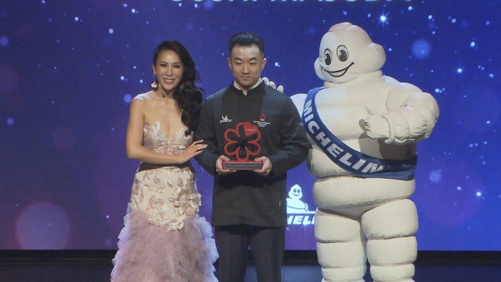 Yoji Masuda stands on stage to accept his award from the presenter and Bibendum, the Michelin Man