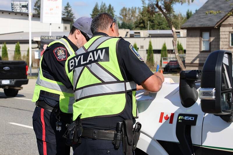 Two bylaw officers write a ticket on the hood of a police vehicle.